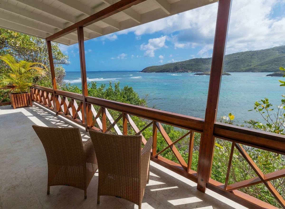 A spacious balcony features two wicker chairs positioned to overlook the serene bay. Lush greenery frames the view, with gently rolling hills in the background under a partly cloudy sky. The wooden railing's design adds charm, enhancing the overall outdoor ambiance.