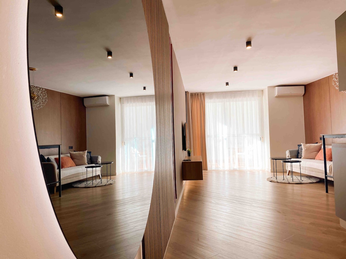 A spacious living area is captured in the reflection of a round mirror. The room features large windows with sheer curtains allowing natural light to fill the space. A minimalist decor is highlighted by a comfortable bed, a side table, and a television unit under softly illuminated ceiling spots.