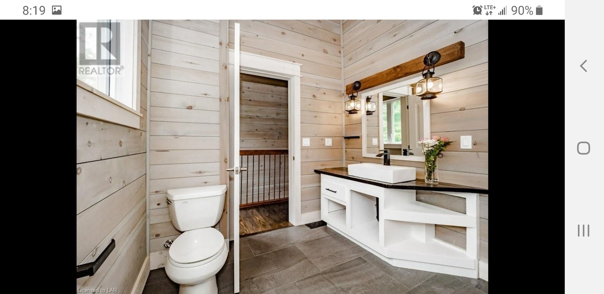 A modern bathroom features a sleek vessel sink atop a white vanity, complemented by dark countertops. Natural wood accents are visible on the wall and light fixtures. The white shiplap walls enhance the space, while the tiled floor provides a neutral foundation.