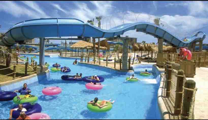 A water park scene features a winding blue waterslide extending over a lazy river. Guests float on colorful inner tubes, with palm trees and shaded lounging areas visible in the background. The sky is clear with a few wispy clouds above, creating a bright, inviting atmosphere.