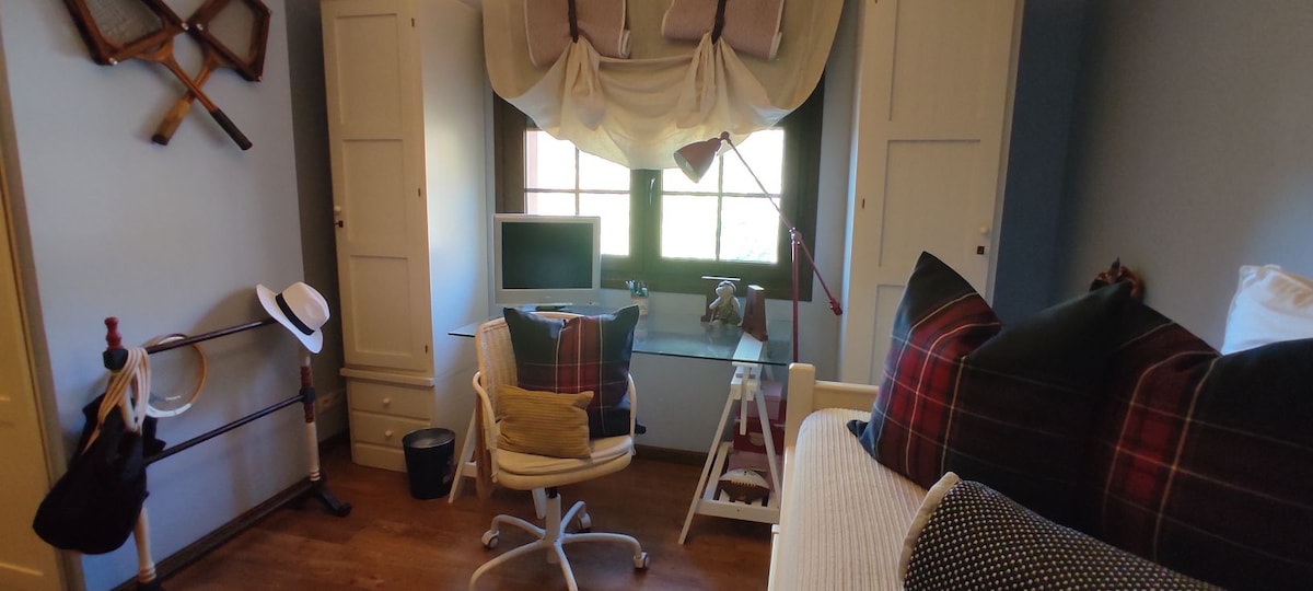 A versatile room features natural light from two large windows, illuminated by a desk lamp. A comfortable chair is positioned near a desk with a computer. Storage is provided by built-in cabinets, while decorative elements include a hat and sporting equipment.