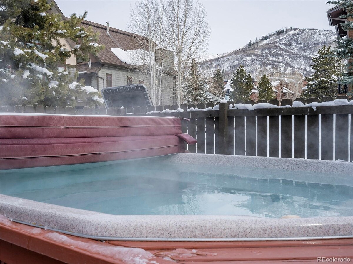 A heated outdoor hot tub is visible, surrounded by a wooden fence and snow-covered ground. Steam rises from the water, indicating a warm and inviting atmosphere against a backdrop of mountains and pine trees.