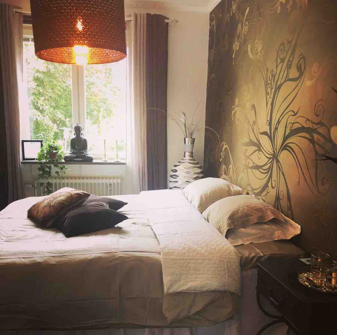 A cozy bedroom features a spacious bed adorned with neutral-toned bedding and decorative pillows. A large window allows natural light to fill the room, complemented by soft drapes. An artistic wall design and a stylish light fixture add to the room's comfort and charm.
