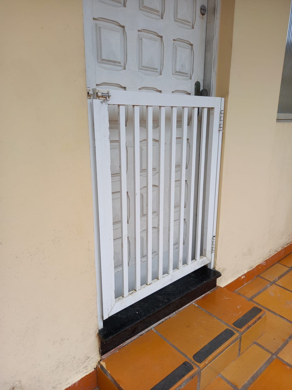 A white security gate is positioned in front of a wooden door, providing an added layer of safety. The gate features vertical bars and is mounted on a painted surface. The surrounding wall is a soft beige, complemented by orange and black tiled flooring.