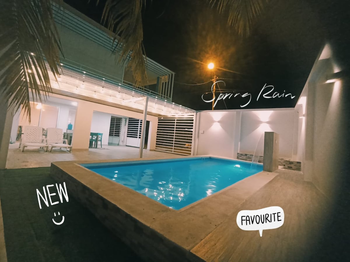 A modern pool area is illuminated in soft lighting during the evening. The pool’s clear water reflects the surrounding structures, while lounge chairs and tables are arranged nearby under a spacious outdoor patio.