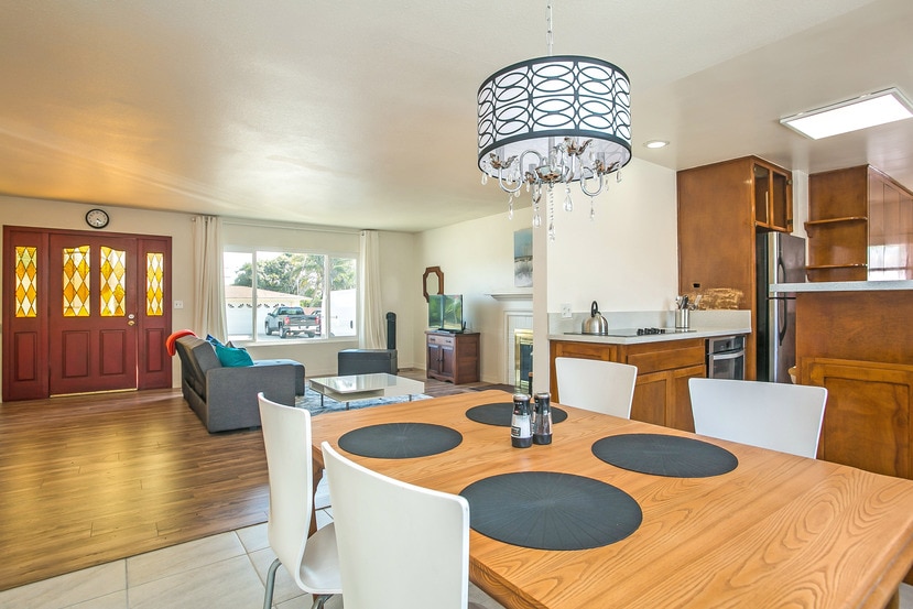 An open and airy living and dining area features a wooden table set for four, with black placemats and a centerpiece. A modern chandelier hangs overhead. Large windows allow natural light to flood in, highlighting a cozy sofa and entertainment unit across the room.
