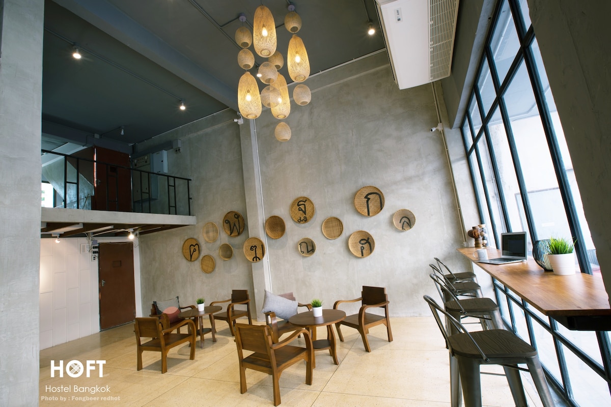 A contemporary common area features large windows allowing natural light to fill the space. Several wooden chairs surround low tables, complemented by decorative circular wall hangings. A seating nook near the windows includes bar stools and long counters for social gatherings.
