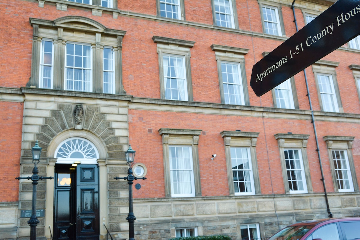 The historic exterior of County House is viewed, showcasing its red brick facade, large windows, and elegant entrance door. A sign marking the apartments is displayed prominently, along with classic lampposts that frame the entrance, highlighting the building's architectural features.