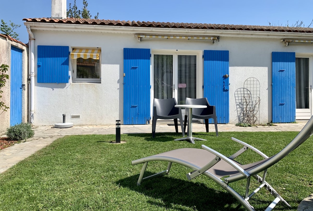 A well-maintained outdoor area is shown, featuring a grassy space with a relaxing lounge chair and a small table with two chairs. The exterior wall of the studio is fitted with bright blue shutters, and a large window is visible, providing a vibrant contrast against the white facade.