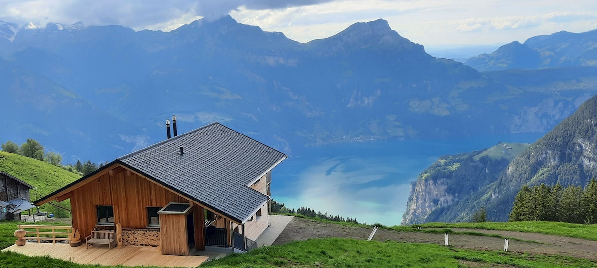 A newly constructed wooden house sits on a hillside, providing panoramic views of the surrounding mountains and a serene lake. The building's alpine style features a combination of natural materials, while a spacious deck invites relaxation in the scenic outdoors.