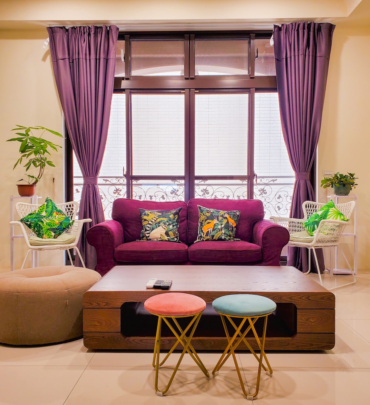 A seating area features a purple sofa adorned with vibrant throw pillows, complemented by two white chairs with green cushions. A wooden coffee table sits in the center, flanked by colorful stools. Natural light filters through large windows draped with dark purple curtains.