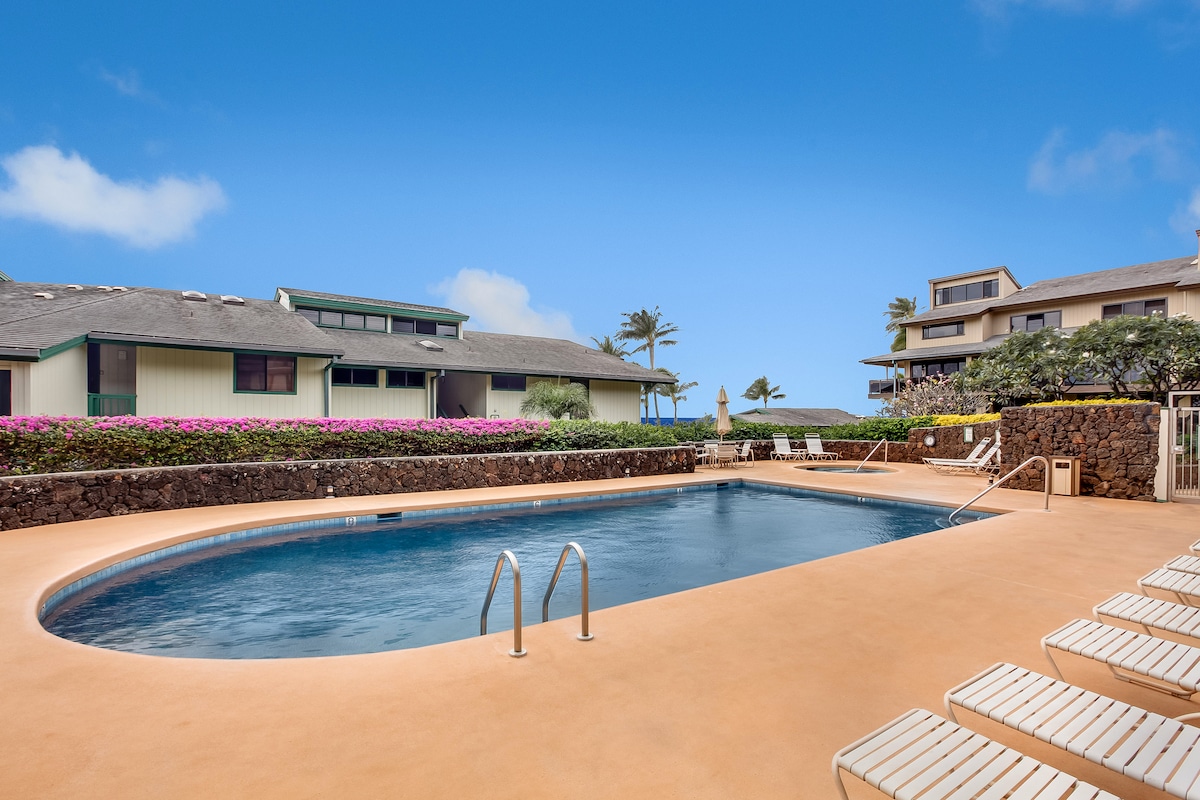 A spacious outdoor pool is framed by a neatly landscaped area featuring colorful flowering plants. Surrounding the pool, cushioned lounge chairs provide ample seating. A few buildings with large windows are visible in the background against a clear blue sky.