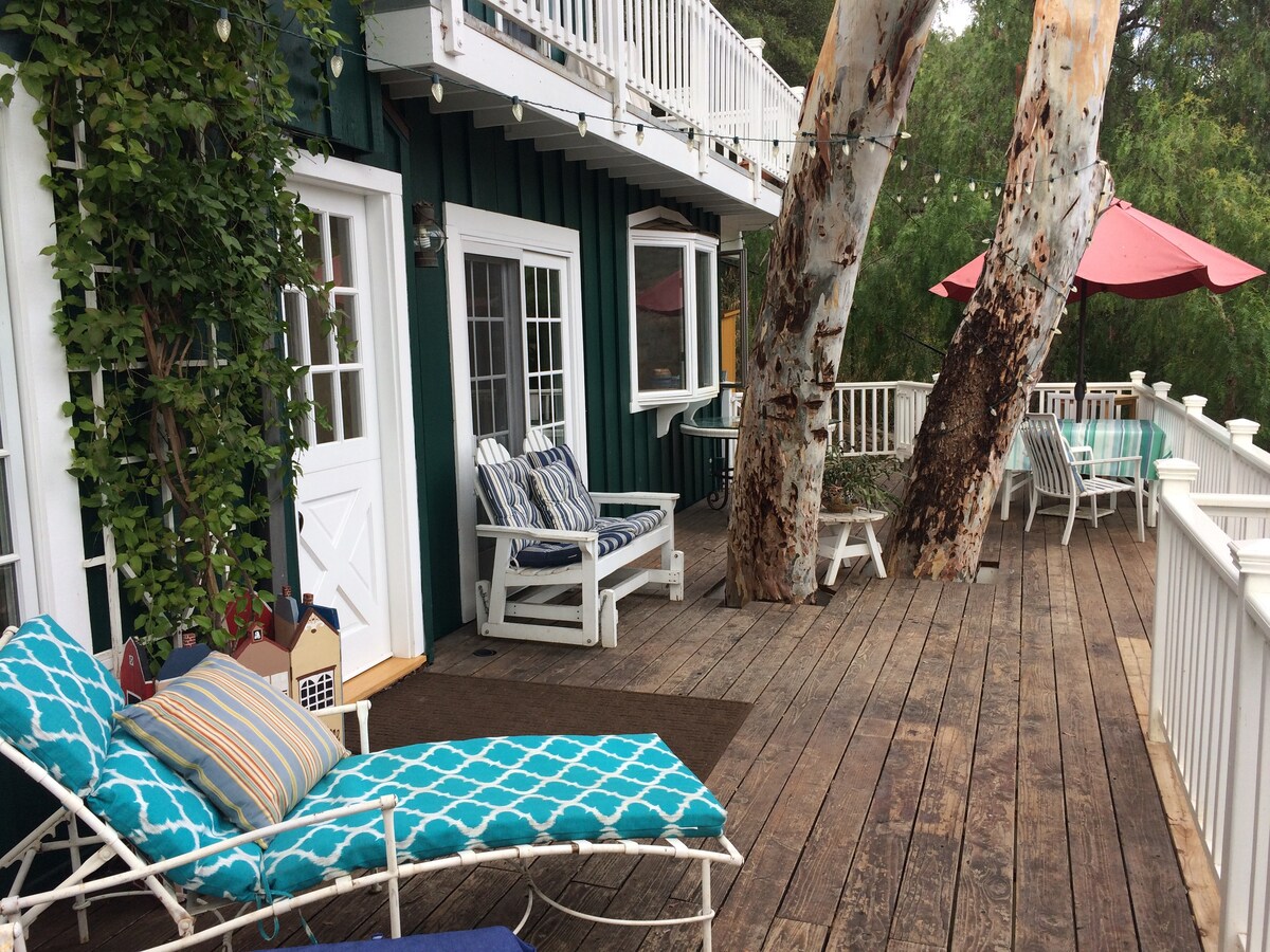 An expansive wood deck incorporates lounging chairs and tables, bordered by greenery and large trees. It features a parasol providing shade and a view of the surrounding landscape, enhancing outdoor relaxation. The deck's design encourages comfortable seating and socializing in a tranquil setting.