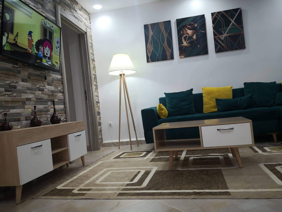 A cozy living area is highlighted by a green sofa adorned with a yellow cushion, complemented by a light wood coffee table. A television mounted on the wall is positioned above a decorative console table, while framed wall art adds visual interest to the inviting space.