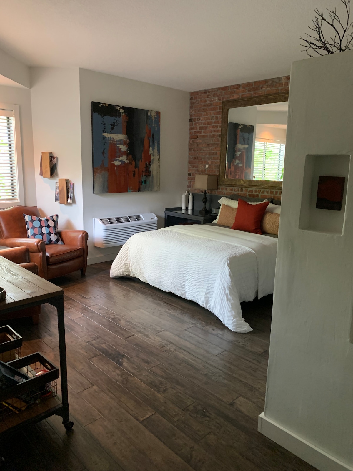 A comfortable guest suite is designed with a spacious bed covered in a white quilt. An accent wall of exposed brick enhances the warm ambiance. A wooden desk and two brown armchairs complement the space, with light filtering through a window dressed with blinds.