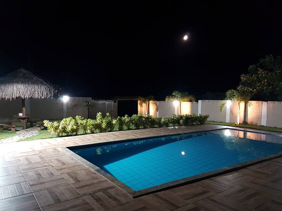 A tranquil pool area is illuminated under the moonlight, with a clear blue pool surrounded by landscaped greenery. A thatched-roof structure offers seating, while soft lighting enhances the peaceful ambiance of the outdoor space.