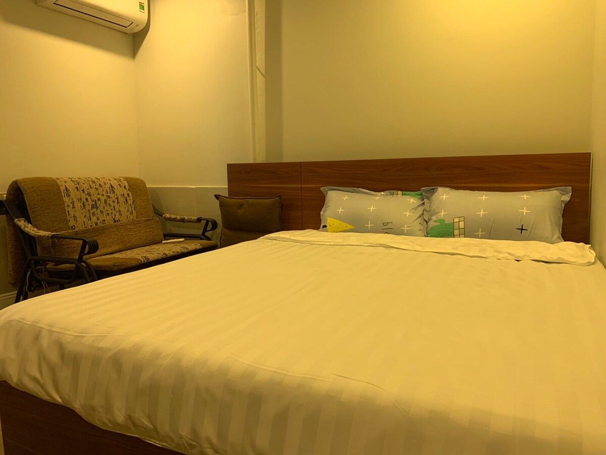 A spacious bed is centered within the room, adorned with light-colored linens. To the side, a cozy seating area features a simple couch and a decorative pillow. Soft, warm lighting creates a calm ambiance, and an air conditioning unit is mounted on the wall above.
