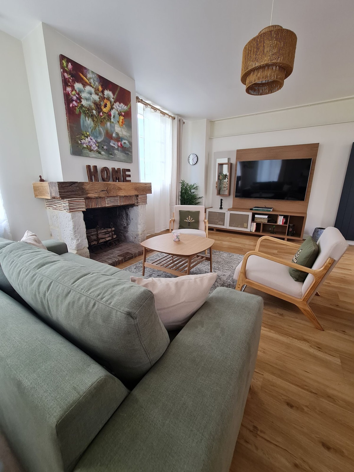 The living area presents a comfortable green sofa alongside a light wooden armchair. A wooden coffee table is positioned in front, accompanied by decorative elements. An artistic floral painting adorns the wall above a rustic fireplace, while a television is mounted on the adjacent wall.