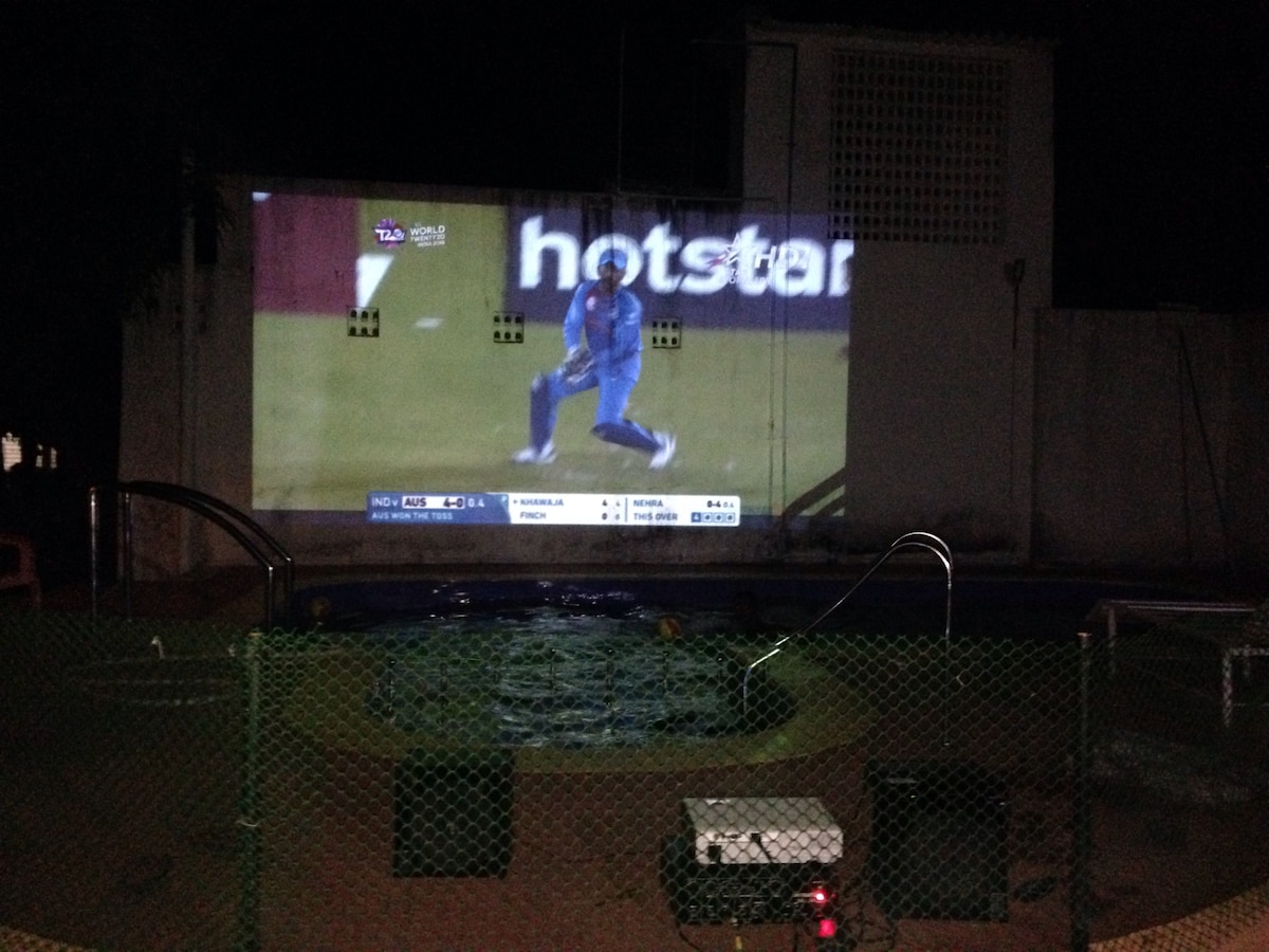 A projector displays a sports event on an outdoor wall, with a swimming pool in the foreground. The pool area is delineated by a green mesh fence, creating a space for relaxation and entertainment. Soft lighting enhances the ambiance of the evening setting.