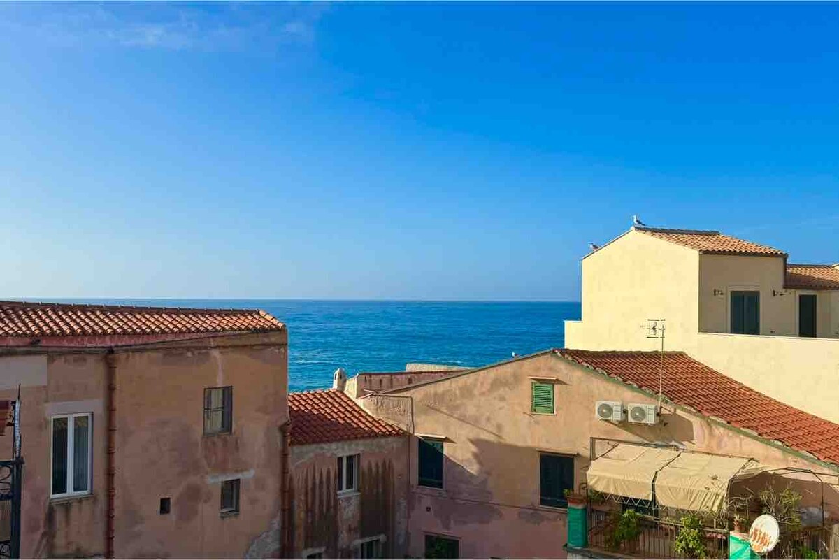 A view of the tranquil sea is captured, showcasing the expansive blue waters against a clear sky. Traditional Sicilian architecture with terracotta roofs is visible in the foreground, creating a harmonious blend of nature and historical buildings.