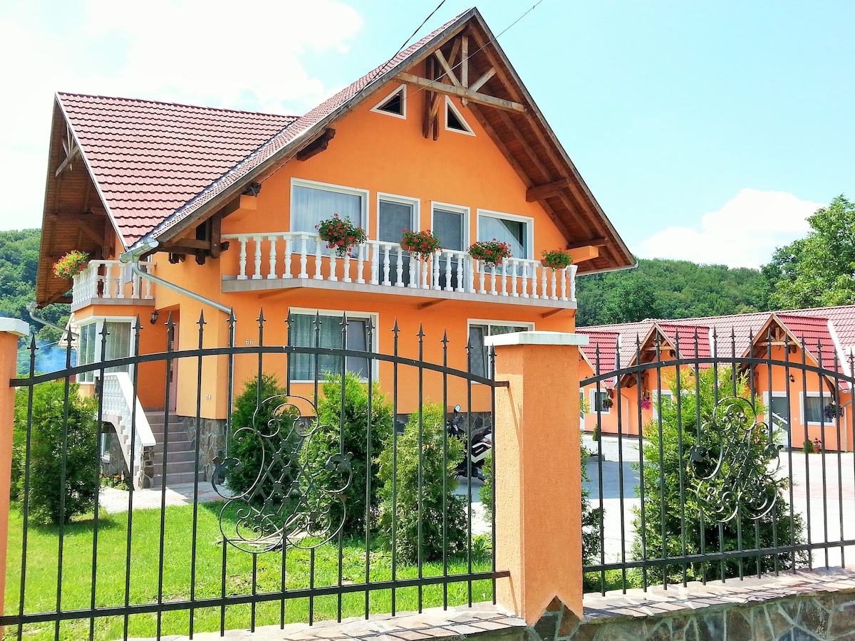The exterior of the house features a warm orange facade with wooden accents and a sloped roof. Flower boxes adorn the windows, and a decorative wrought iron fence encloses the property, which is surrounded by green lawns and shrubbery.