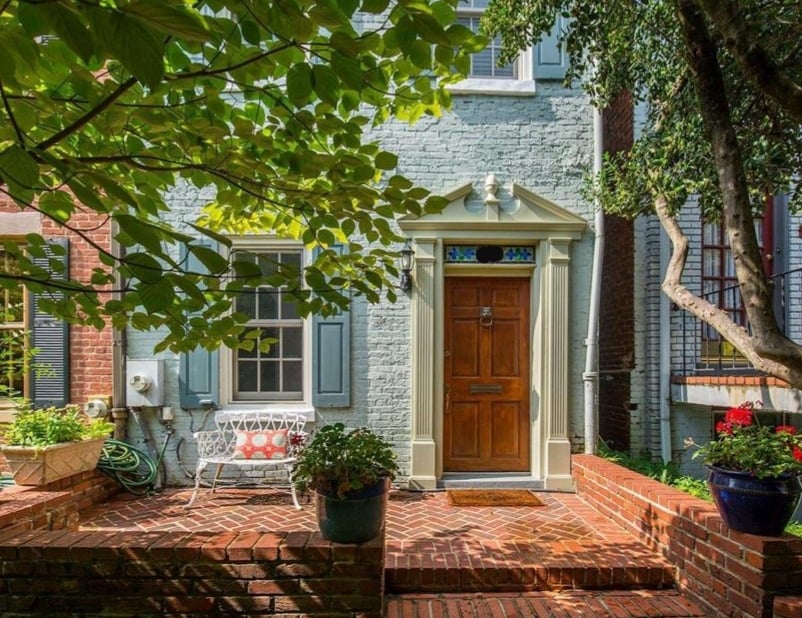 The entrance features a simple wooden door framed by a decorative arch. Lush green foliage and potted plants create a welcoming atmosphere. A brick pathway leads to the entrance, with a small bench providing a cozy sitting area in front of the house.