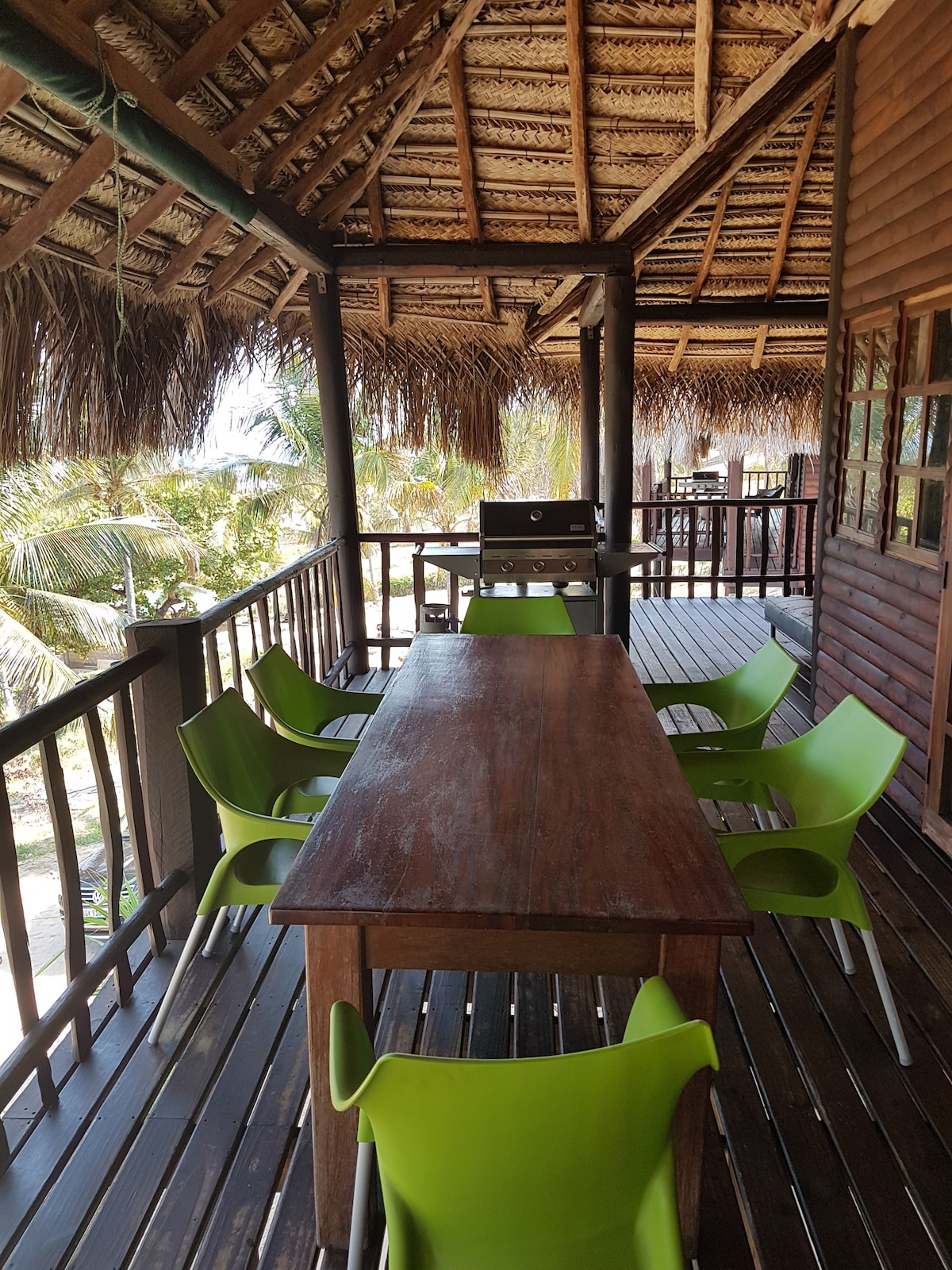 A spacious outdoor dining area features a wooden table accompanied by bright green chairs. The open space is covered by a thatched roof, with views of surrounding palm trees visible in the background. A barbecue grill is positioned nearby for cooking.