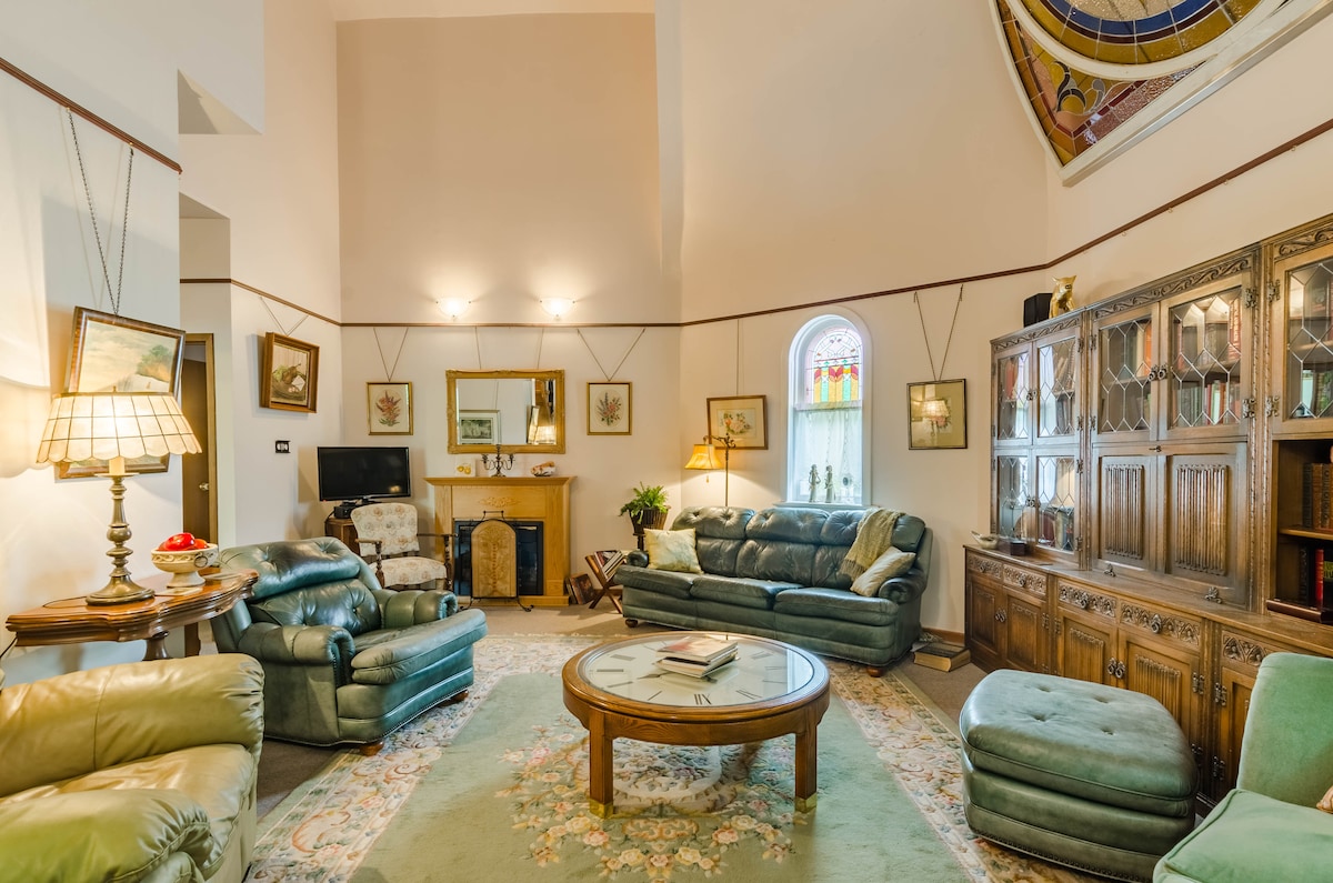 A spacious living area features comfortable leather seating arranged around a central table. Soft lighting is provided by twin lamps, and a cozy ambiance is complemented by decorative art on the walls and a wooden cabinet filled with books. A beautiful stained glass window enhances the space.