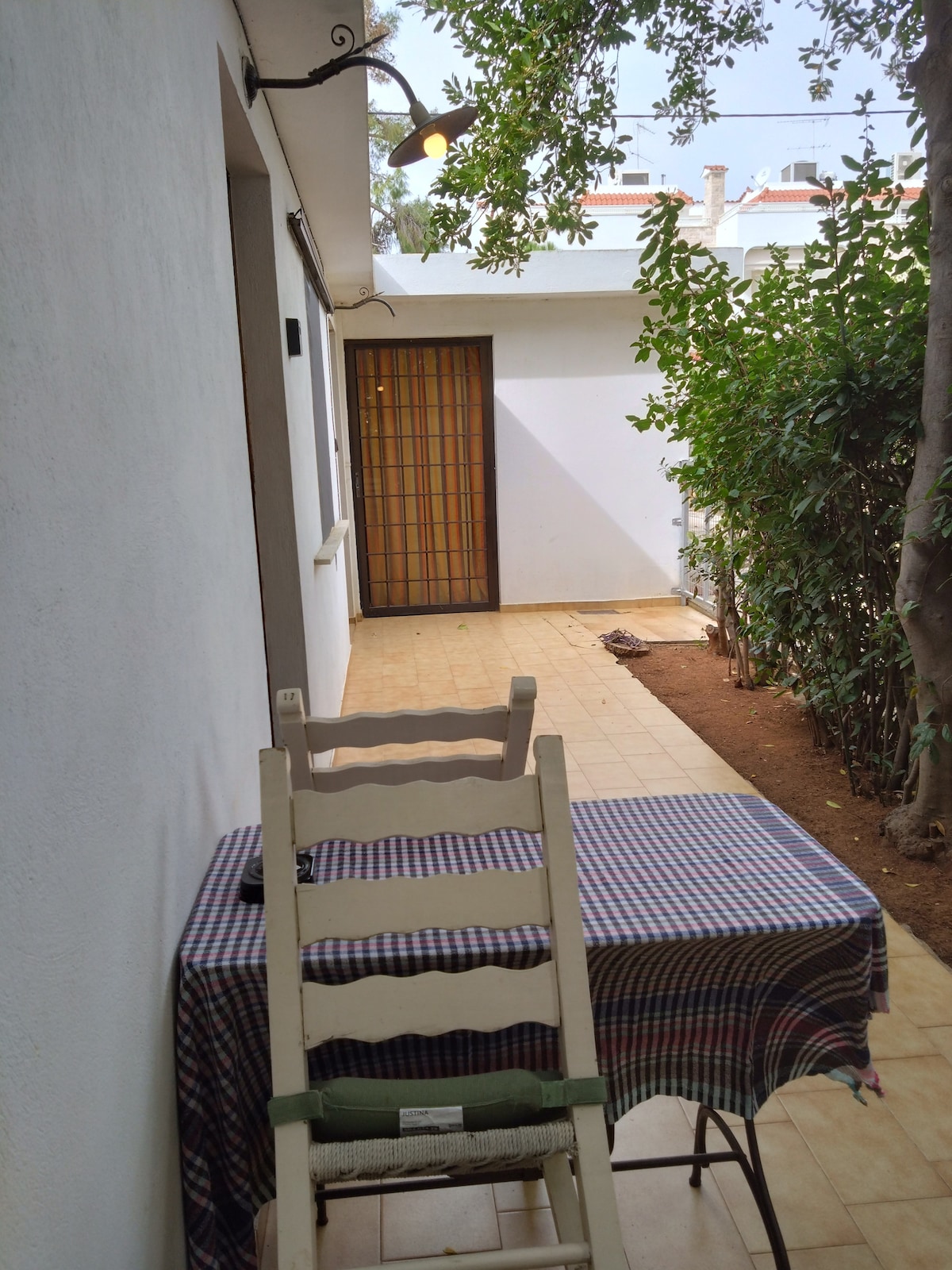 A private patio area is shown, featuring a traditional checkered tablecloth draped over a small table. A chair is positioned at the table, complementing the serene garden view. Lush greenery borders the space, and ample natural light is visible through the entrance.