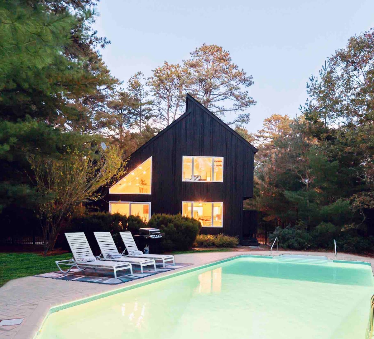 A modern Scandinavian home is depicted, surrounded by trees. Large windows reflect warm light, creating a welcoming glow. A rectangular pool, bordered by lounge chairs, offers a relaxing outdoor space. The structure features a distinctive angled roofline, enhancing its contemporary design.