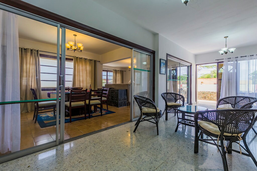 An open space connects the living and dining areas, framed by large glass doors. Rattan chairs with light cushions surround a glass table, creating a casual seating arrangement. Natural light filters through sheer curtains, enhancing the airy feel of the environment.