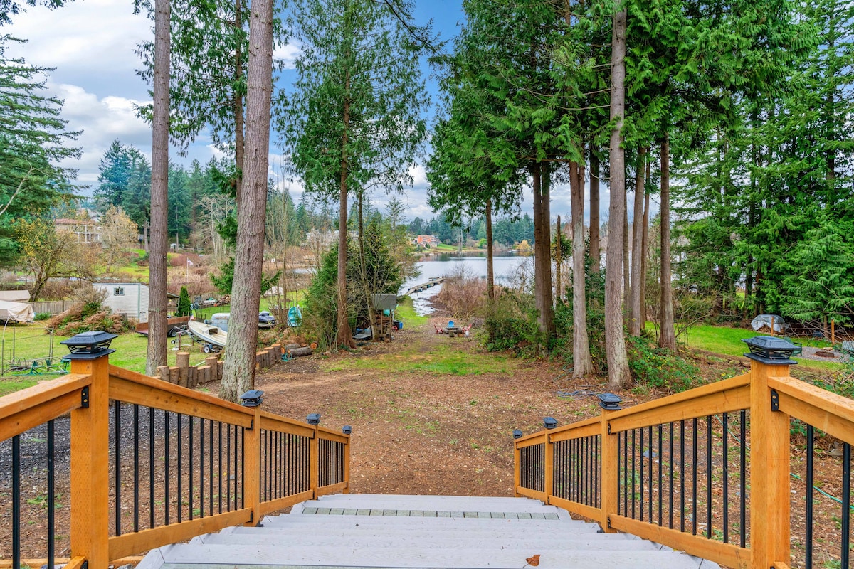 A wooden staircase leads down to a spacious, grassy area surrounded by tall trees. A serene view of the lake is visible in the background, with fishing boats dotting the shoreline. Natural light filters through the leaves, enhancing the peaceful outdoor setting.