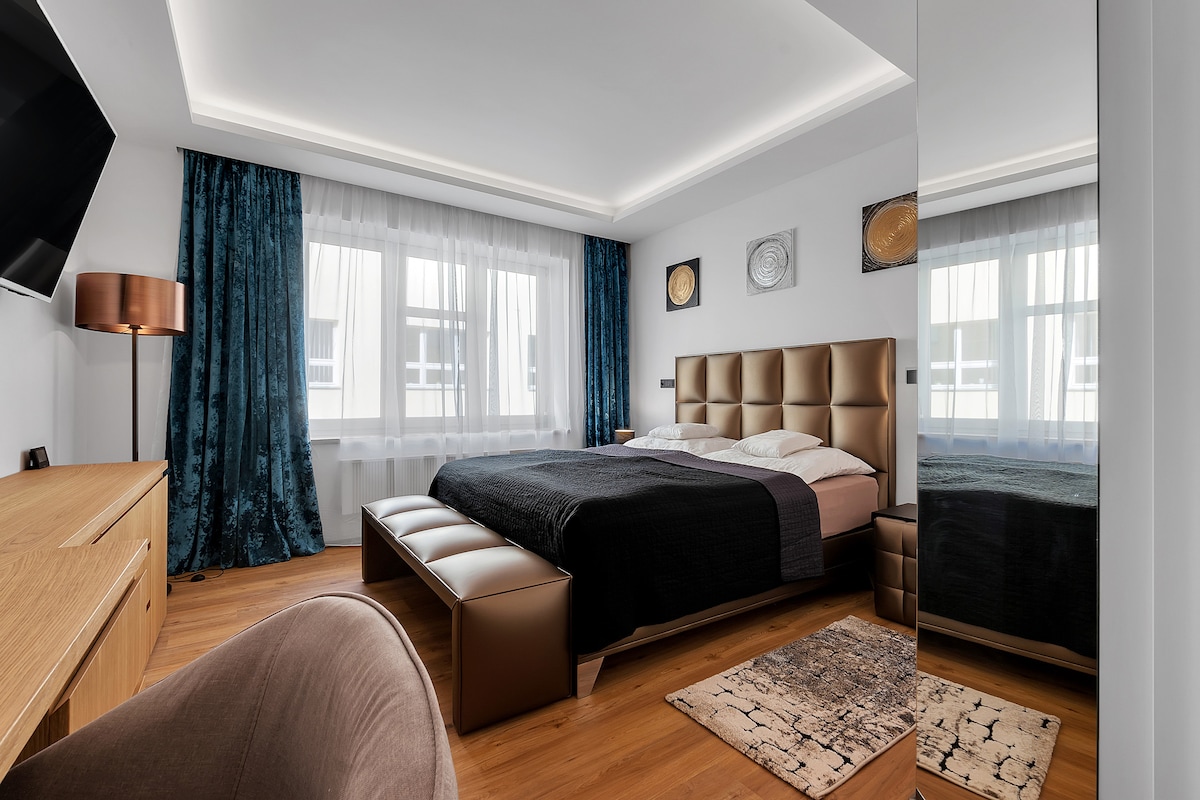 A well-appointed bedroom features a spacious bed with an elegant headboard, complemented by a stylish bench at the foot. Large windows dressed in sheer curtains invite natural light, while a mirrored wardrobe enhances the room's dimensions. A subtle area rug adds warmth to the wooden floor.