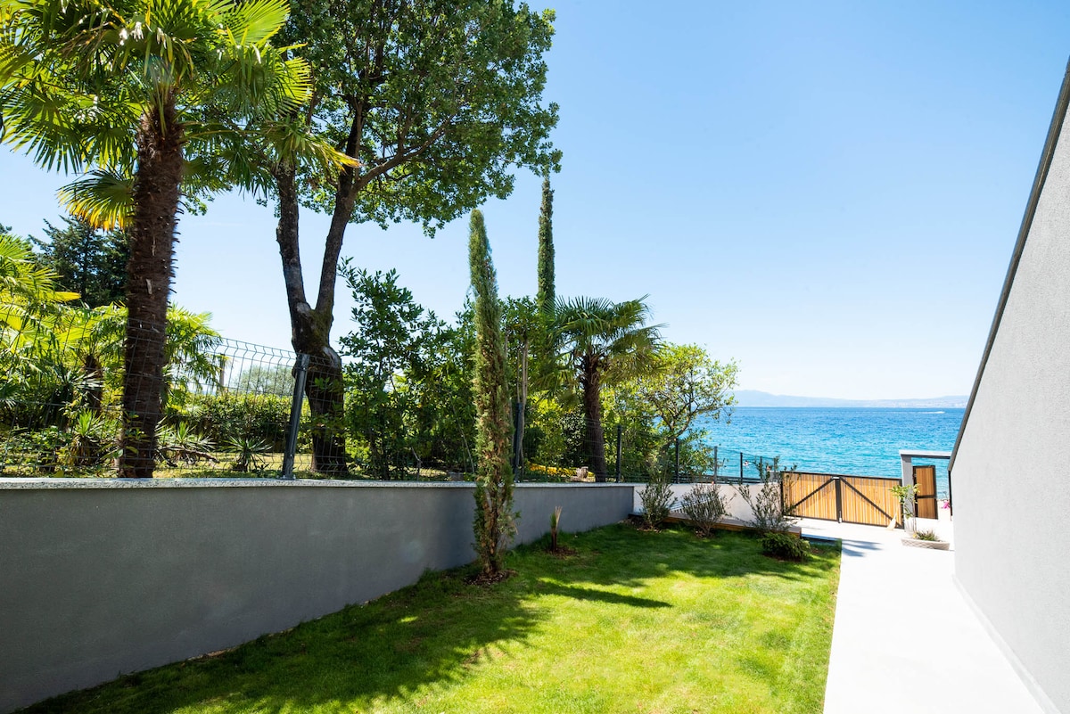 A green lawn is visible, bordered by tropical plants and trees. In the background, the glistening sea can be seen beyond a small gate. The clear blue sky complements the serene setting, creating a peaceful outdoor space.