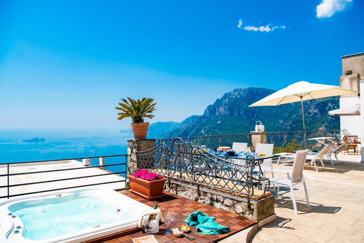 An outdoor terrace is showcased with a heated Jacuzzi and sun loungers. A large umbrella provides shade, while a potted plant and colorful flowers add a touch of greenery. The backdrop features expansive views of the mountains and coastline under a clear blue sky.