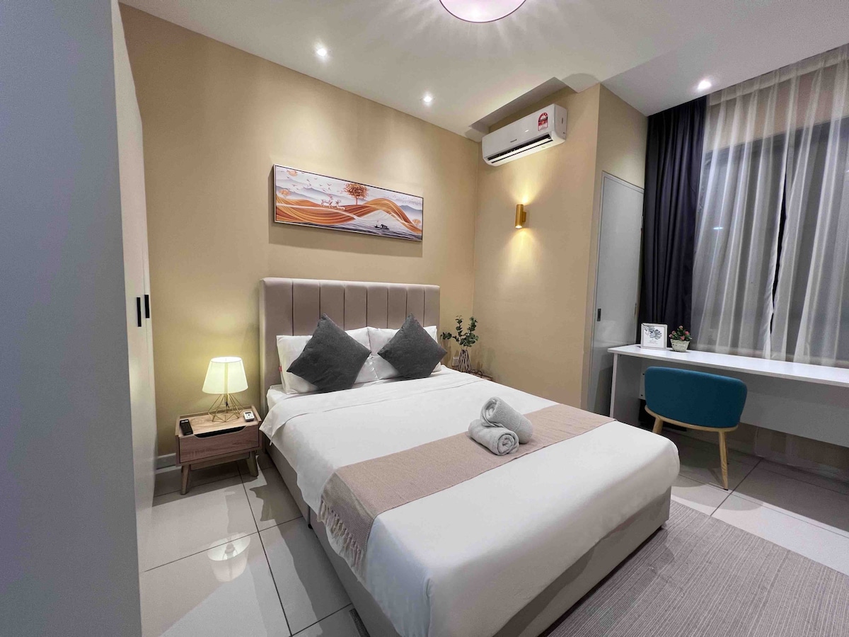 A softly lit bedroom features a spacious bed with neutral bedding and two decorative pillows. A small bedside lamp illuminates the corner, while a desk with a blue chair and a window draped with sheer curtains contribute to the overall modern design.