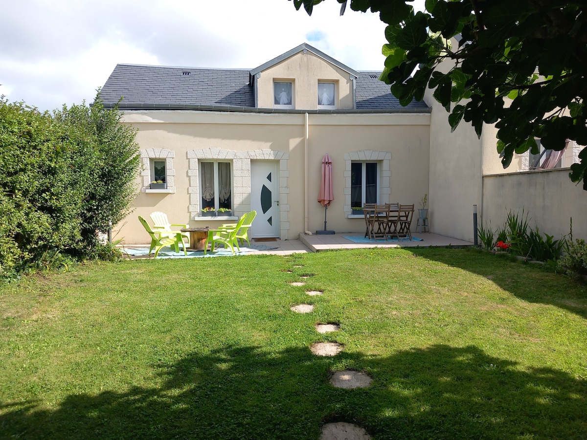 A spacious garden is bordered by neatly trimmed greenery. The patio area features a table with chairs and lounge seating, complemented by a large umbrella. The residence showcases multiple windows, with a light-colored facade and a distinctive door design visible.