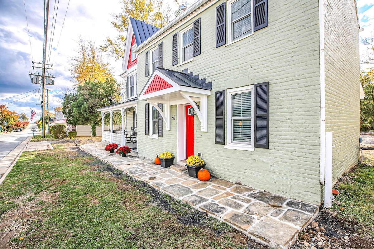 The exterior of the townhome features a charming brick facade in light green, accented by dark shutters. A vibrant red door stands as a focal point, surrounded by welcoming flower pots with seasonal blooms. The covered porch provides a cozy entryway, and the nearby street is lined with trees.