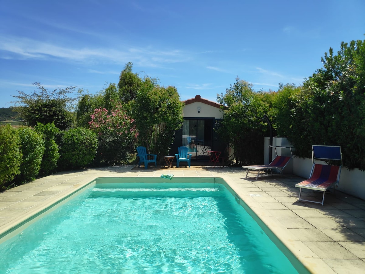 A clear blue pool is surrounded by a tiled patio, featuring bright green shrubs and flowering plants. Two lounge chairs and a pair of blue chairs are positioned near the water, with a small table visible in the background alongside a garden structure.