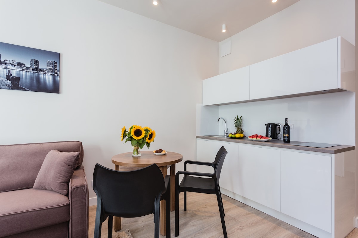 An open-plan living area features a small round table set for two, complemented by a vase of sunflowers. A compact kitchen area showcases modern appliances, including a kettle and a fruit platter. A comfortable sofa is positioned nearby, enhancing the inviting space.