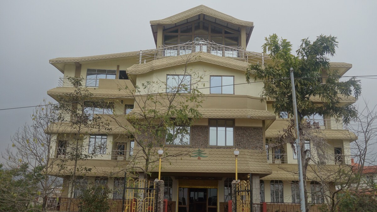 The hotel exterior showcases a multi-level structure with a distinctive roof design and large windows. Natural materials are incorporated into the facade, complemented by surrounding greenery and a welcoming entrance framed by decorative pillars and soft lighting.