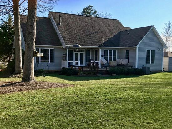 A contemporary home is set in a lush green yard, showcasing a spacious deck with seating and a shaded area. The exterior features a blend of light-colored siding, a pitched roof, and a large window arrangement, inviting abundant natural light into the interior.