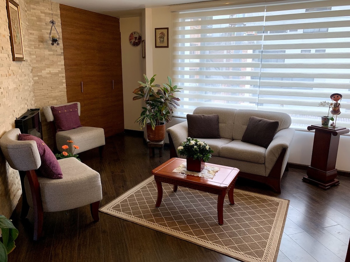 A cozy living room features a plush gray sofa and two armchairs arranged around a polished wooden coffee table. A decorative rug lies beneath, complementing a potted plant positioned beside the window, which is covered by light-filtering blinds that diffuse natural light.