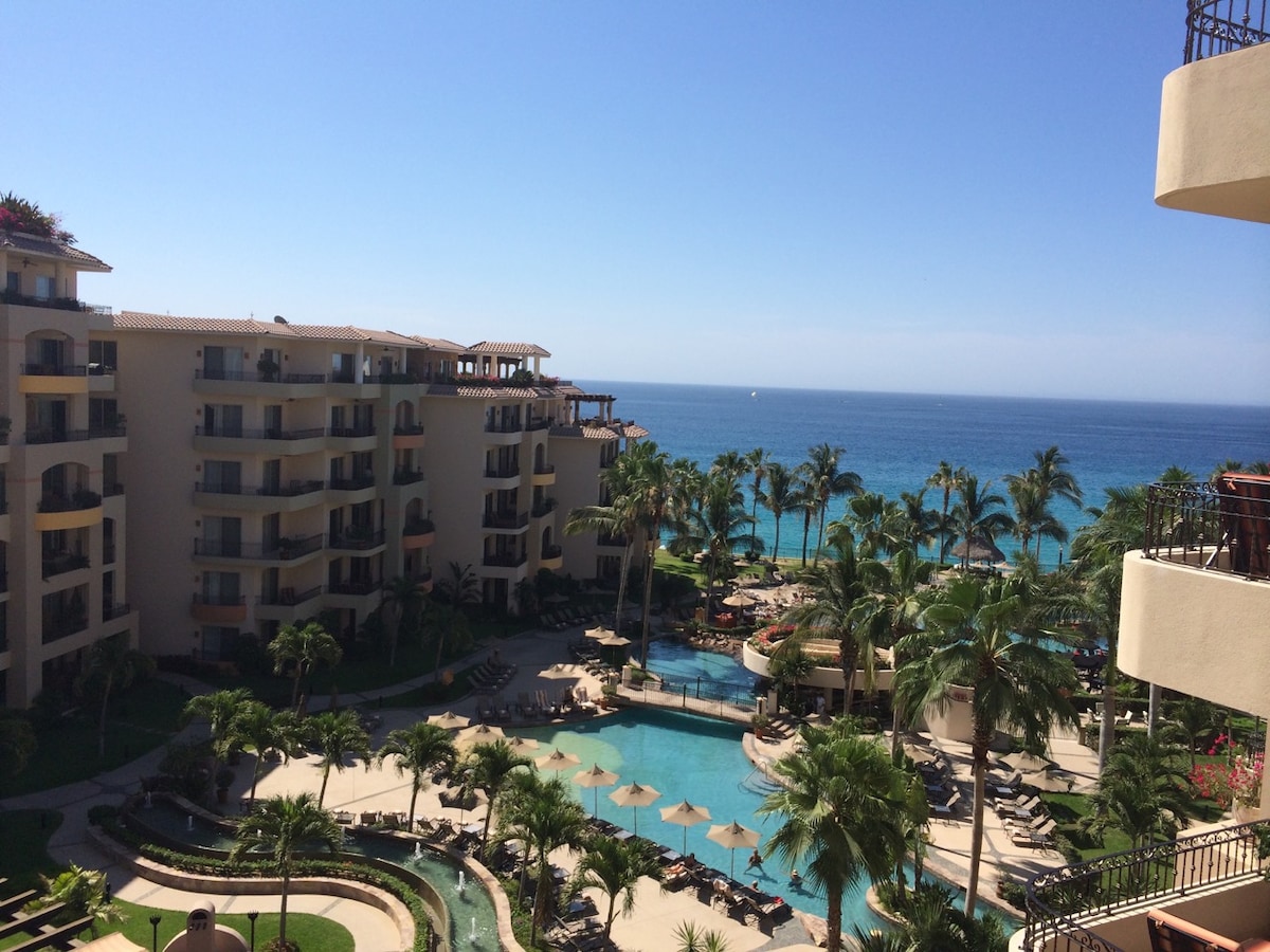 A panoramic view captures the stunning coastline and vibrant pool area. Lush palm trees frame the image, while lounge chairs are arranged around the water. The resort buildings reflect a harmonious architectural style against the clear blue sky and ocean.