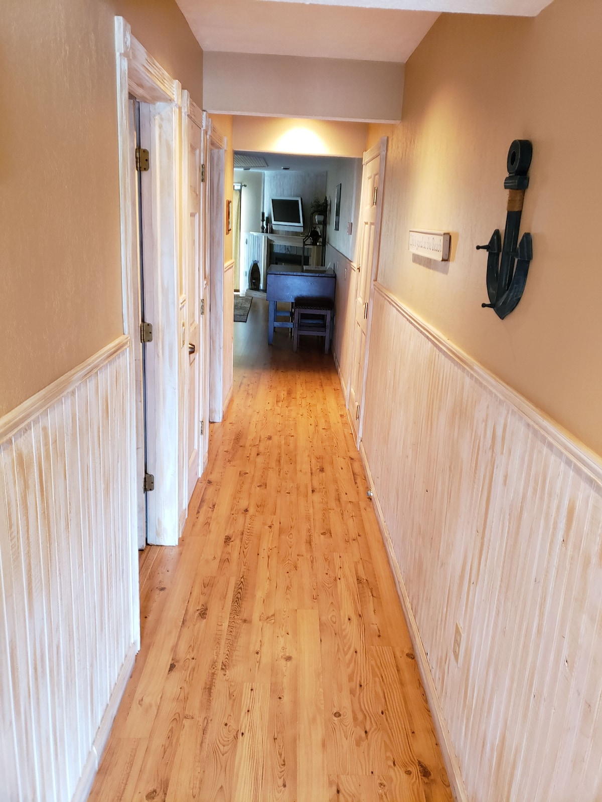 A well-lit hallway features wooden floors and a light beige color scheme. The walls are decorated with a white wainscoting and a wall-mounted anchor. Doors are visible on both sides, leading to various rooms within the condo.