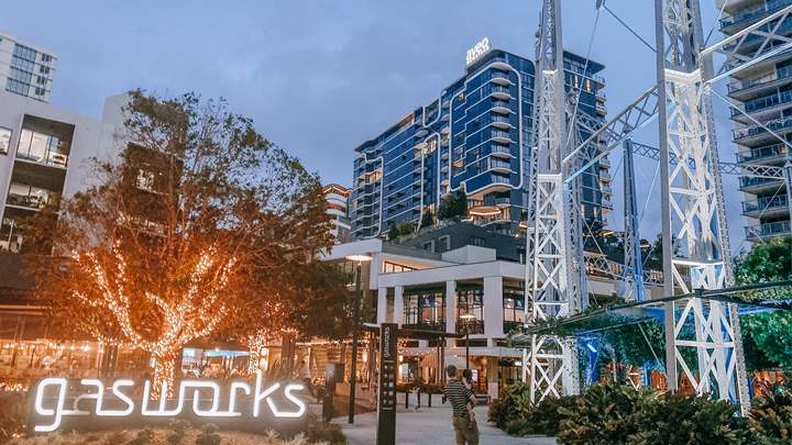 An urban area is illuminated by soft lighting, featuring a large tree with lights and a modern building with a welcoming atmosphere. A bridge structure is visible, reflecting the blend of contemporary design with vibrant nightlife. The scene captures the lively spirit of the Gasworks precinct.