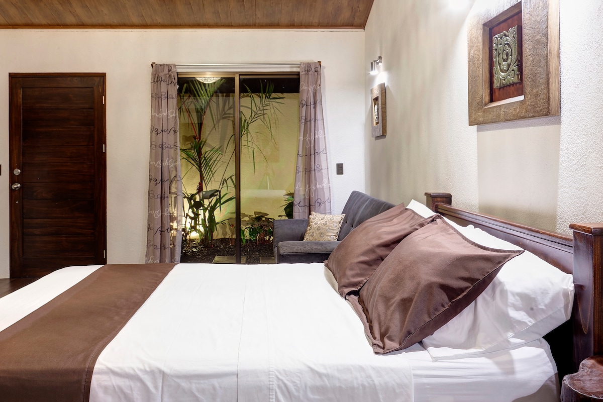 A spacious sleeping area features a large bed with white linens and dark brown pillows. A cozy seating area includes a sofa in the background, while glass doors open to a small garden filled with lush plants, allowing natural light to fill the room.