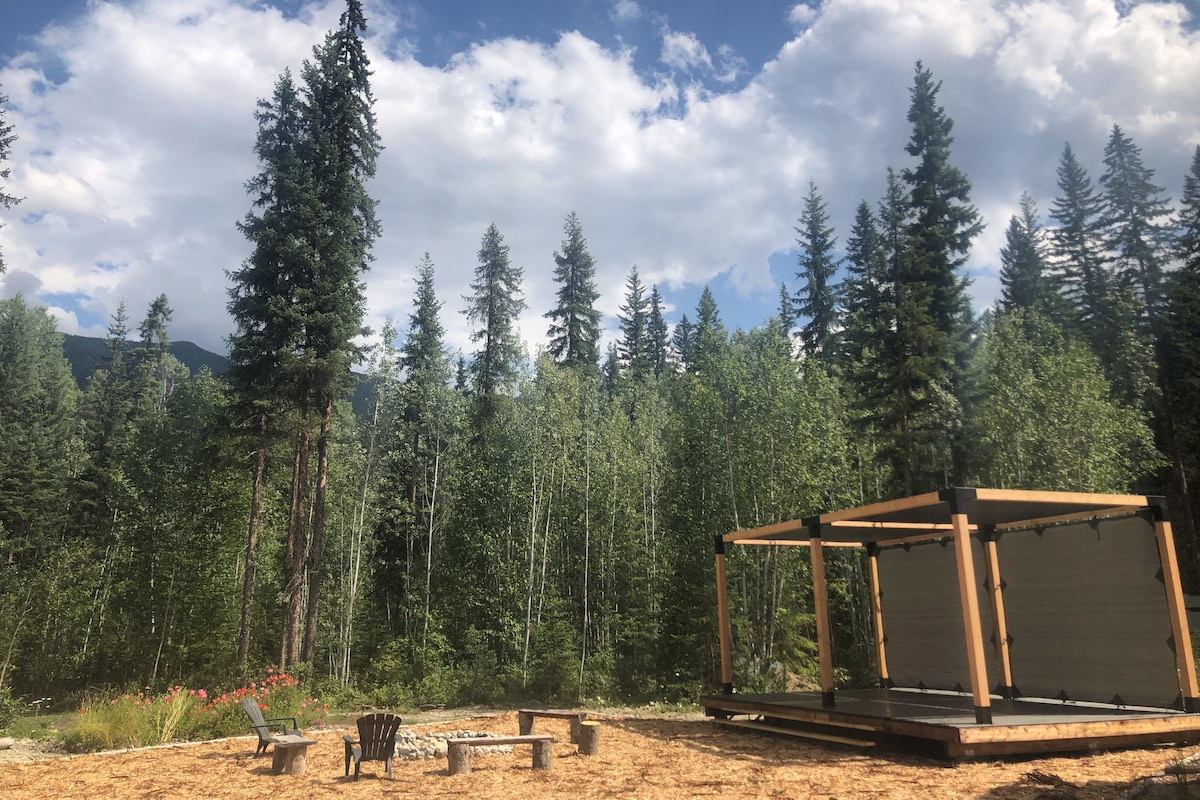An outdoor space is presented, featuring a fire pit surrounded by four adirondack chairs. A partially enclosed structure is located nearby, with lush trees and greenery providing a natural backdrop against a bright sky, enhancing the serene atmosphere.