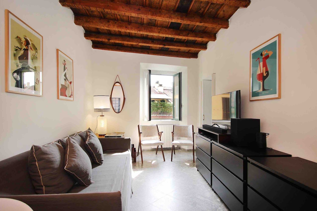 A living area displays wooden beam ceilings complemented by light-colored walls. A gray sofa with decorative pillows faces a sleek black media console beneath a wall-mounted television. Two armchairs provide additional seating near a window, allowing natural light to fill the space.