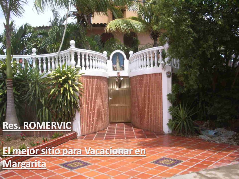 A welcoming entrance features a large, ornate wooden gate framed by lush greenery and tropical plants. The pathway is bordered by a bright tiled area, enhancing the inviting atmosphere of the space.